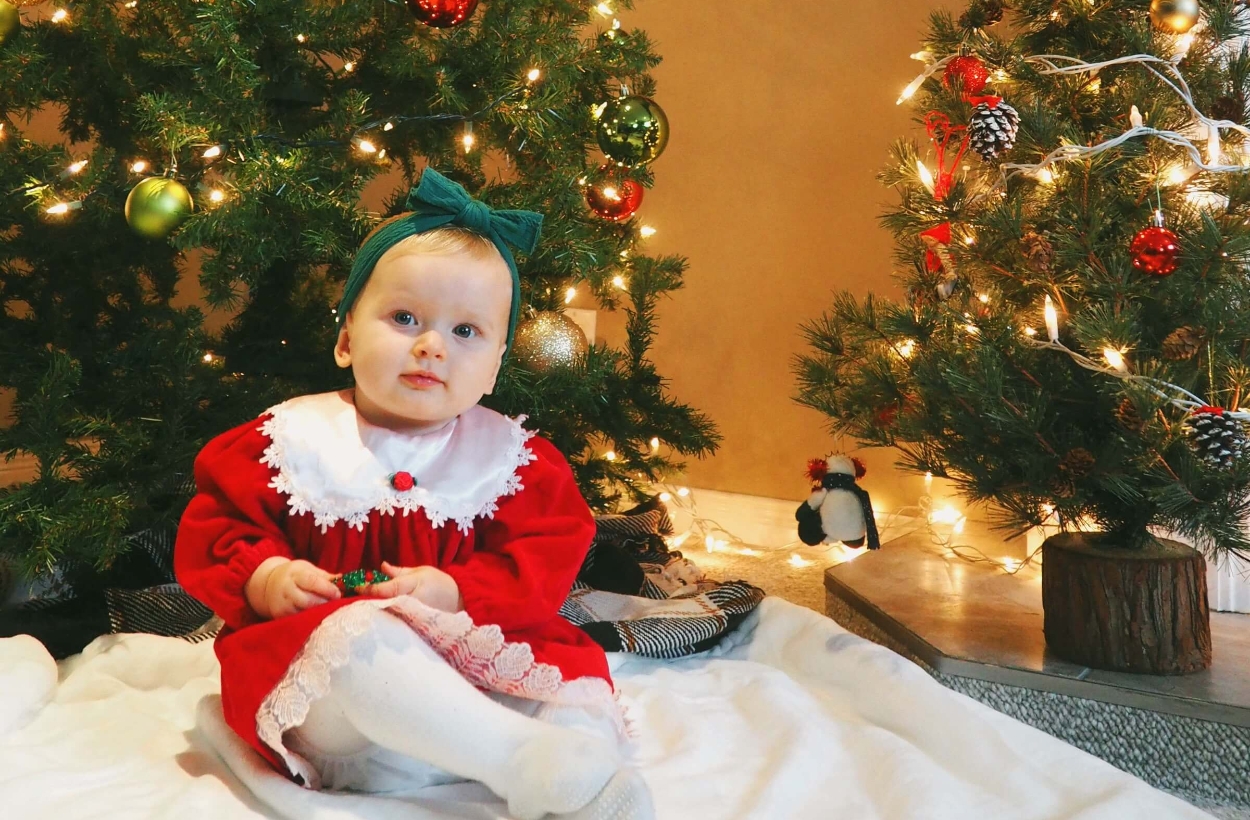 A baby in a red dress sitting in front of the Christmas tree