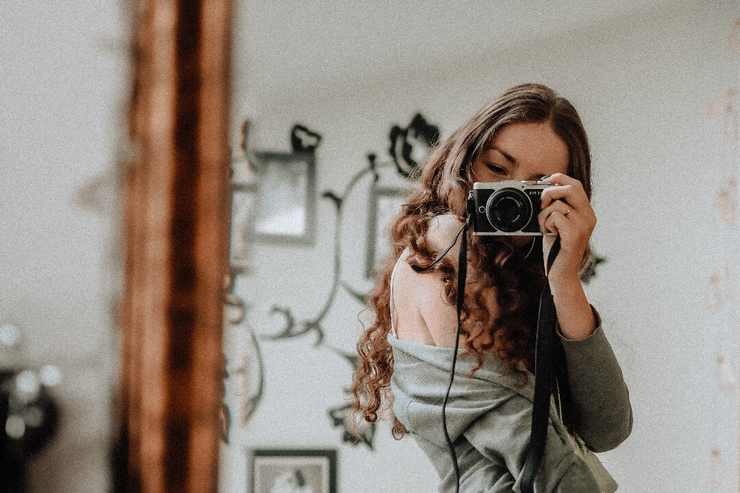 A girl with curly hair is taking a selfie with a camera in front of the mirror