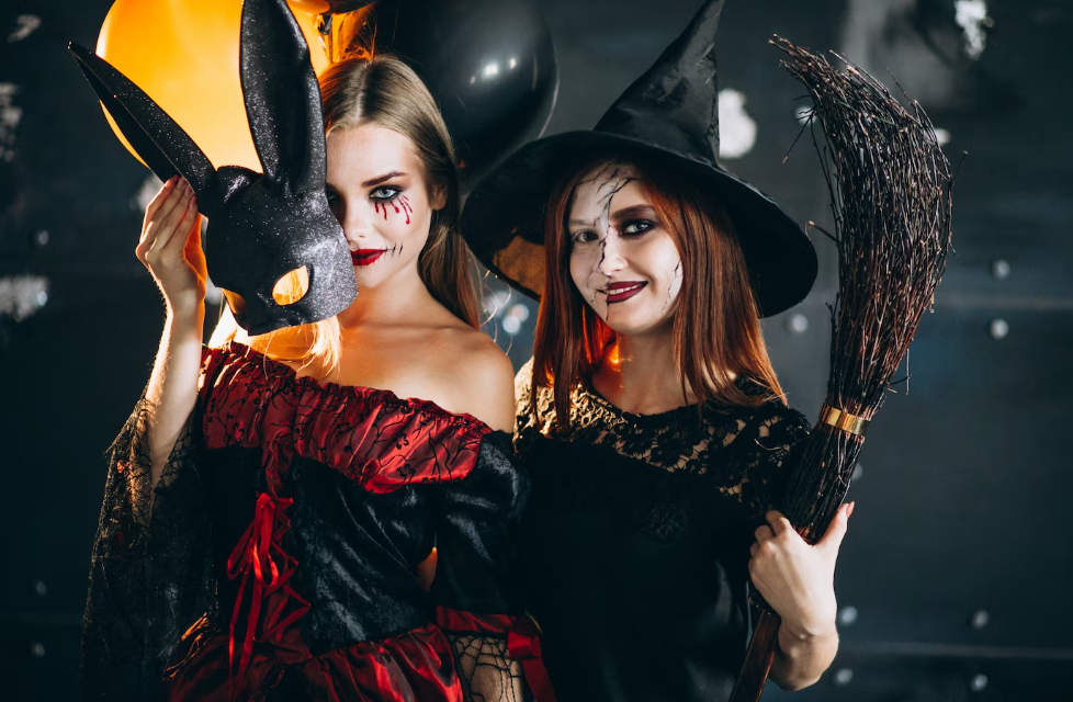Two women in witch makeup, one holding a broom, the other a mask