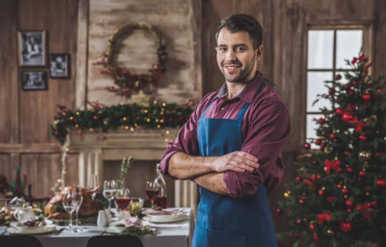 a smiling man in a blue apron stands inside a house with Christmas decors